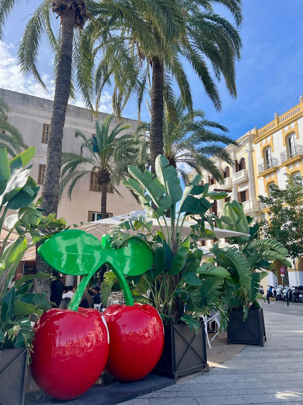 Decorative red cherries with green leaves on a stand in an outdoor setting with palm trees and buildings.