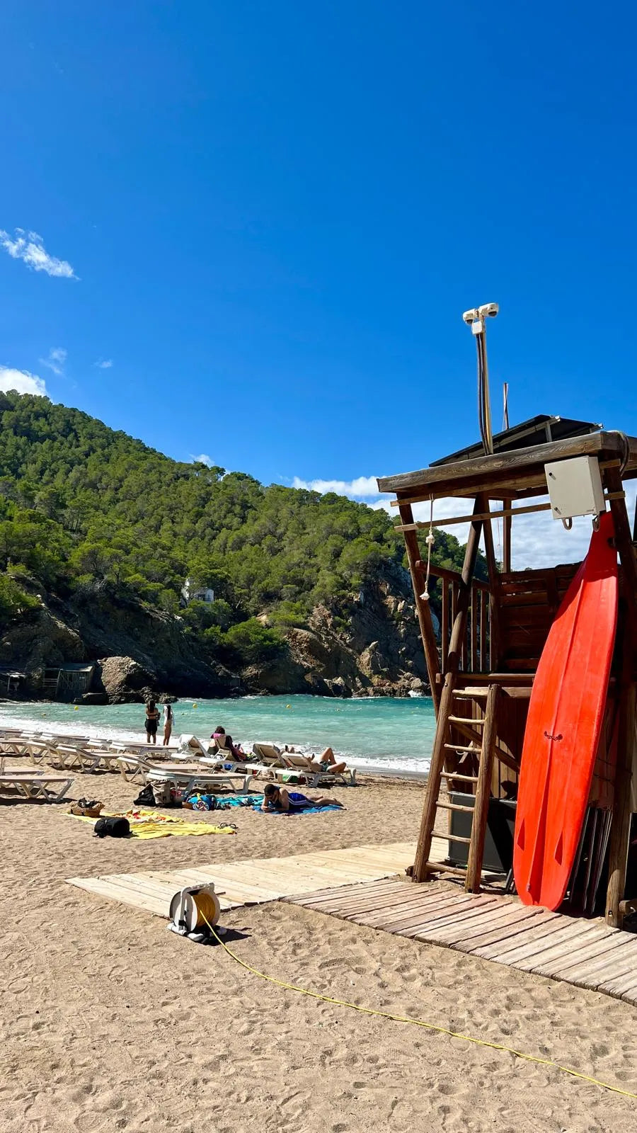 Beach scene with a lifeguard tower, red kayak, and people in the water under a clear blue sky.