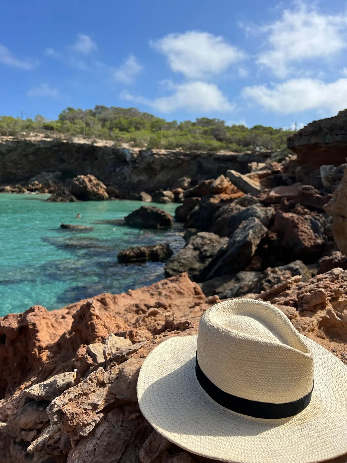 Straw hat with black band on rocky coastal landscape with turquoise water and blue sky.