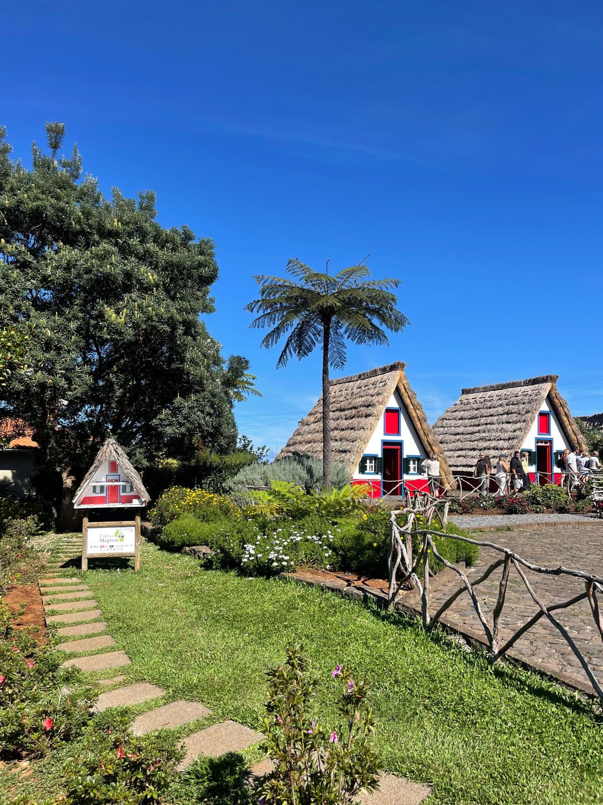 Three thatched-roof cabins with red doors in a garden setting under a clear blue sky.