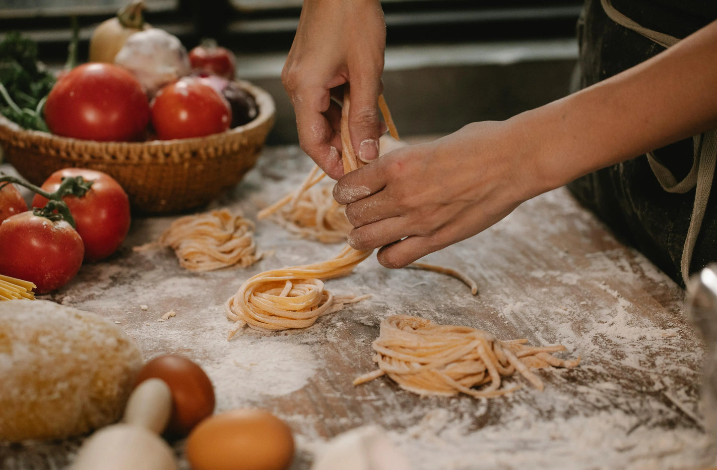 Person making pasta on a wooden table with tomatoes and bread in the background
