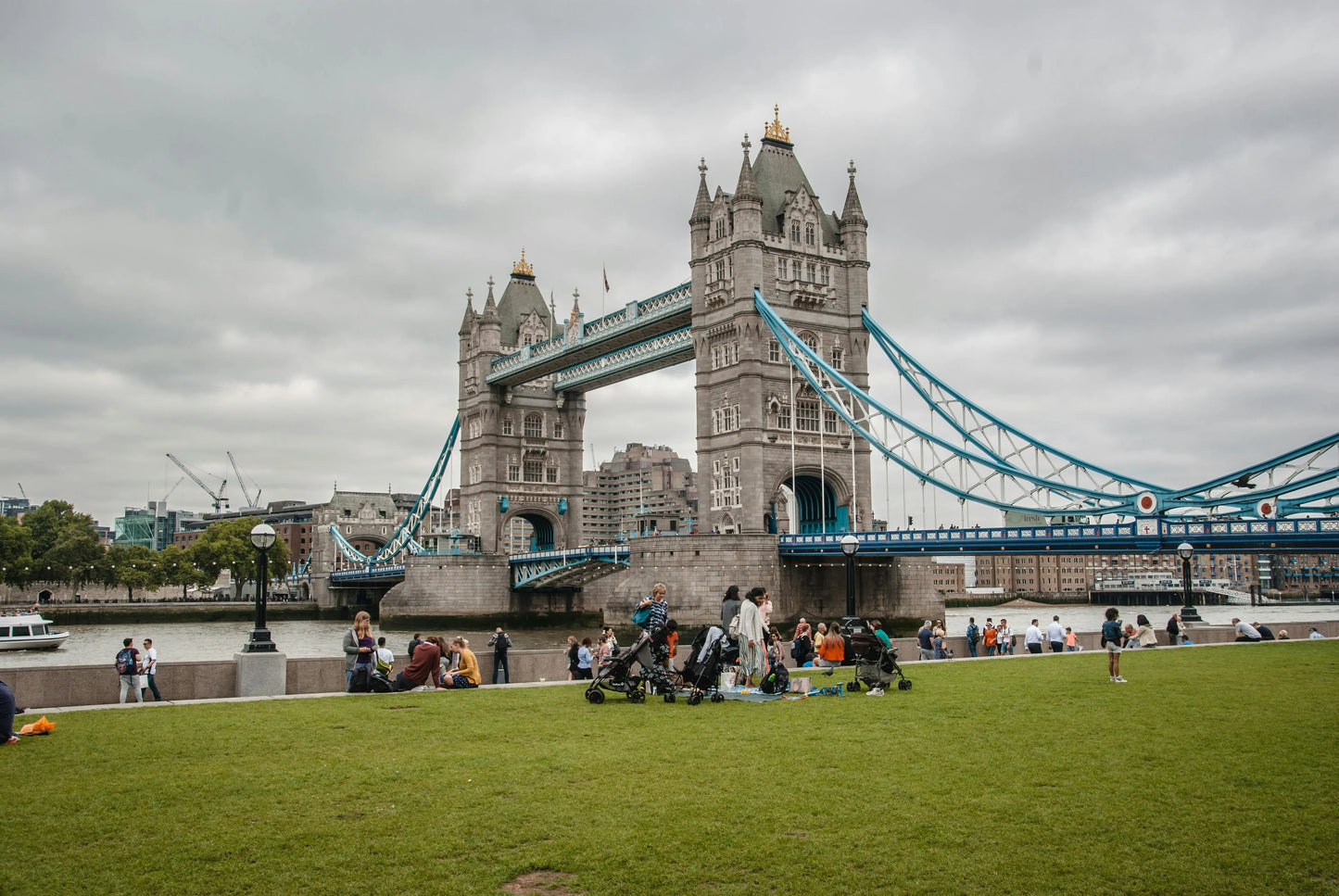 People sitting on a grassy area near Tower Bridge on a cloudy day.