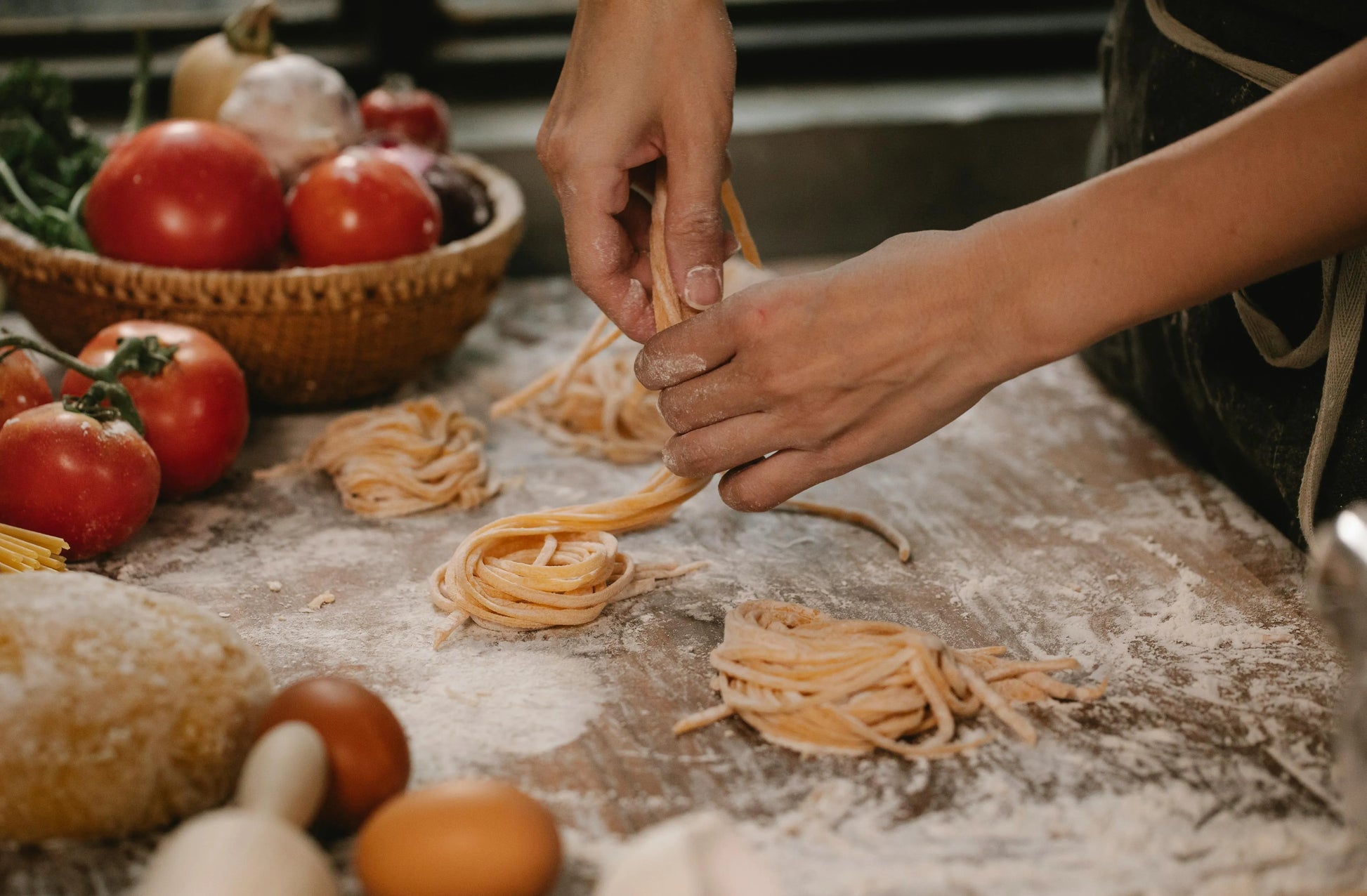 Person making pasta on a wooden table with tomatoes and bread in the background