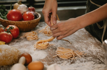 Person making pasta on a wooden table with tomatoes and bread in the background